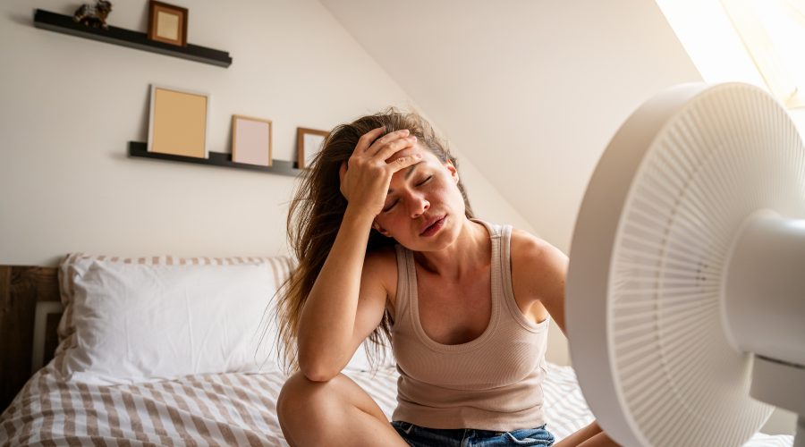 Woman sitting in a hot bedroom using a fan, struggling with uneven cooling in home.