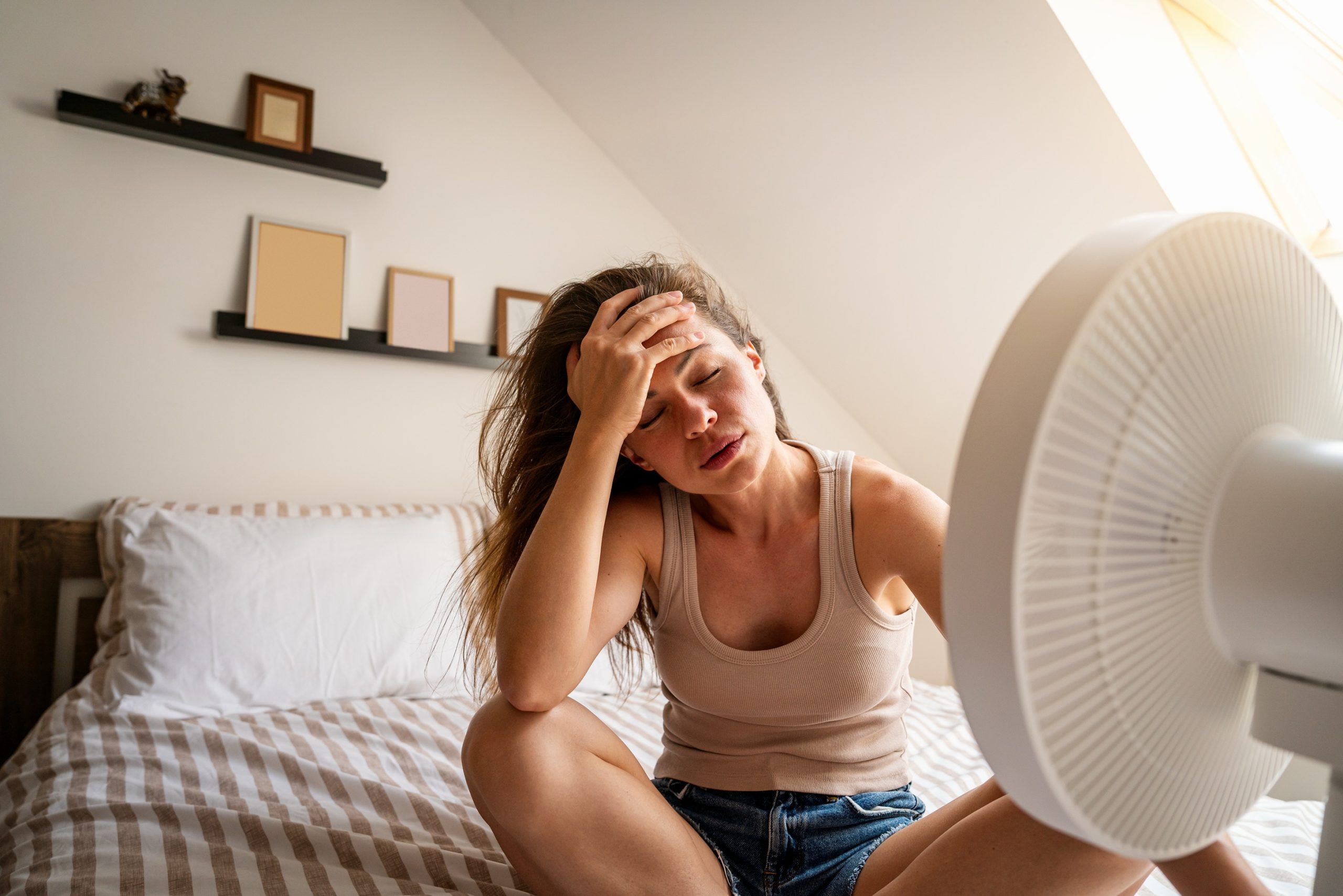 Woman sitting in a hot bedroom using a fan, struggling with uneven cooling in home.